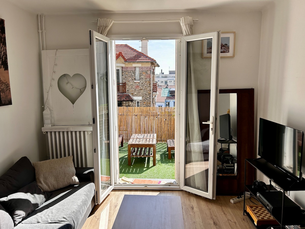 A living room is shown with glass doors opening to a private terrace. Natural light fills the space, highlighting a cozy gray sofa and a TV unit. A mirror reflects parts of the room, while outdoor furniture can be seen through the doors.