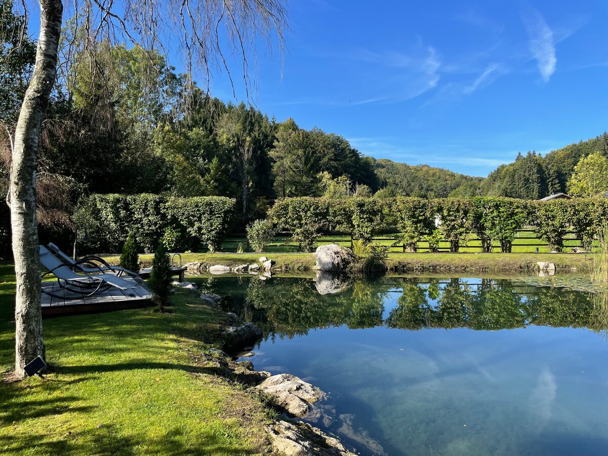 A serene garden scene features a large swimming pond surrounded by lush greenery and trees. The water reflects the blue sky and distant hills. Comfortable loungers can be seen on the grass, offering a peaceful spot to relax by the water's edge.