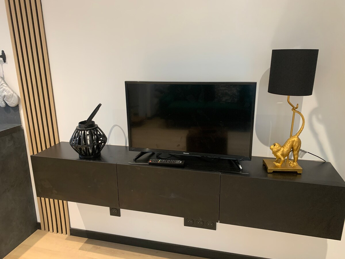 A sleek wall-mounted entertainment unit holds a flat-screen television, accompanied by a decorative lamp with a golden lion figure. A black vase with intricate detailing adds texture to the minimalist setup against a clean, light wall.