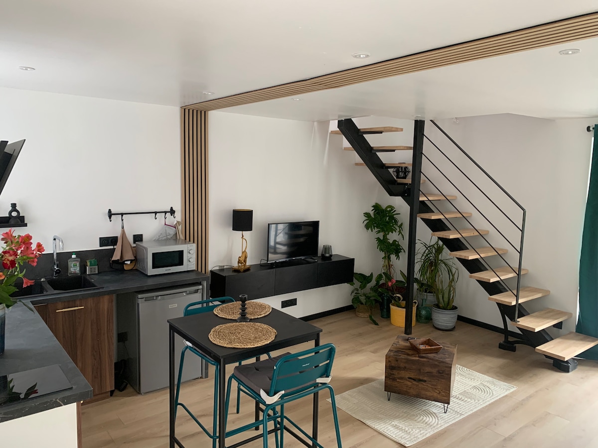 The living area showcases an open layout with modern decor. A kitchen area is equipped with essential appliances, while a dining table with chairs is situated nearby. A wooden staircase leads to the upper level, and potted plants add a touch of greenery to the space.