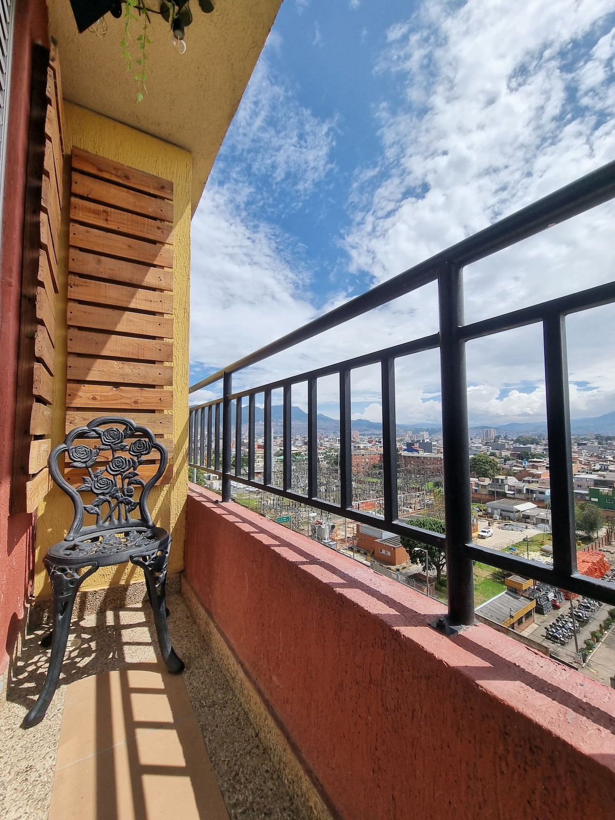 A balcony is presented with a decorative metal chair against a backdrop of open skies and city views. The balcony railing features vertical bars, allowing an unobstructed view of the landscape below, which includes buildings and greenery.