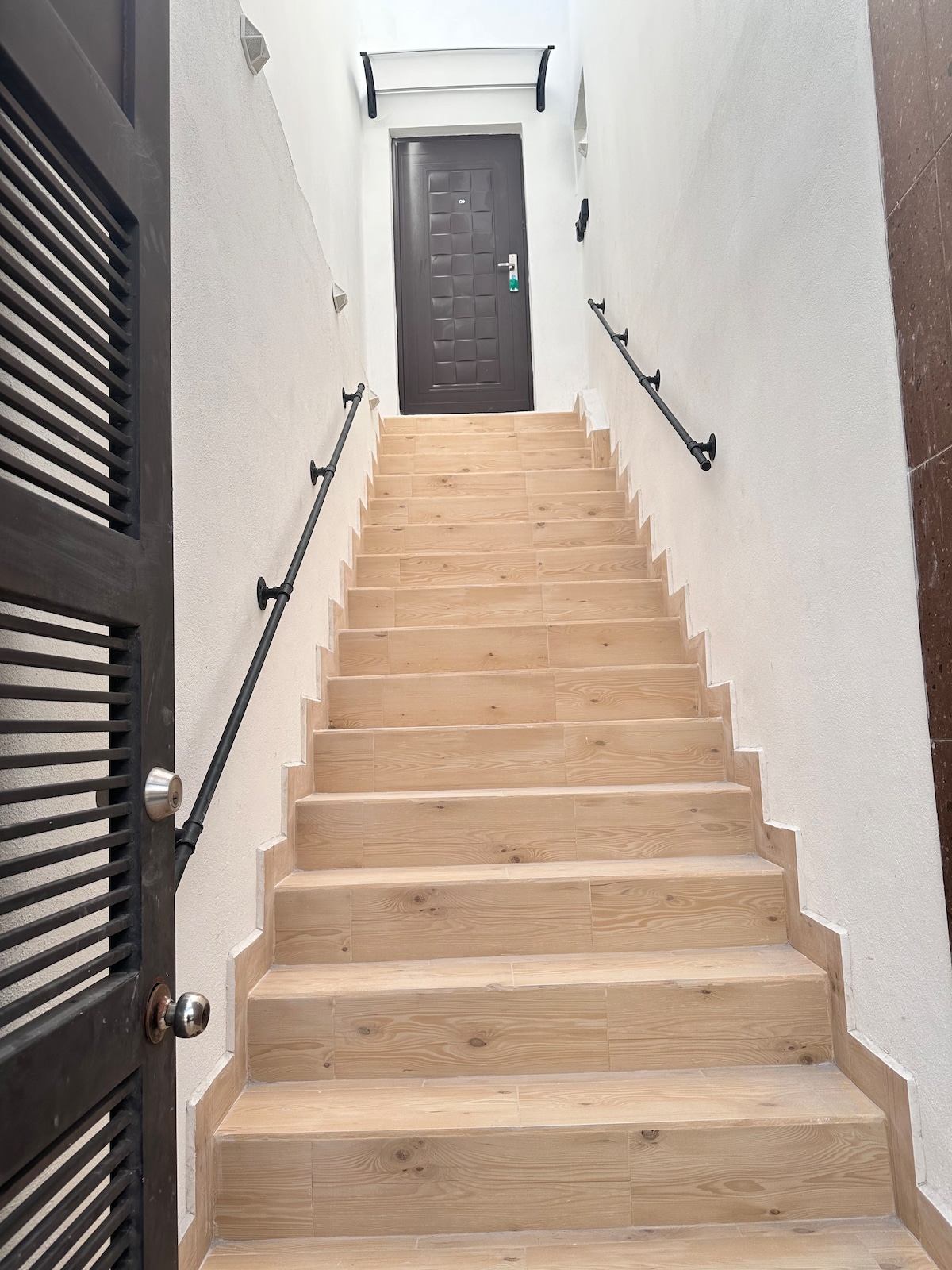 A well-lit stairway features wooden steps leading to a door at the top, with black handrails on either side for support. The white walls create a clean and open atmosphere.