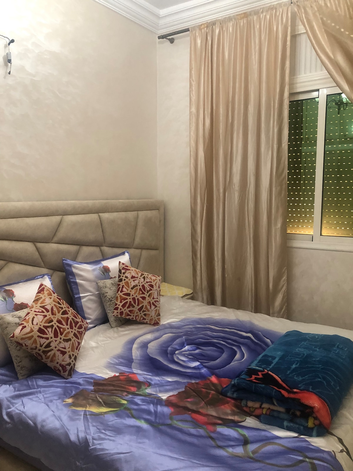 A serene bedroom features a plush bed adorned with a colorful floral bedspread and decorative pillows. Soft curtains frame the window, allowing natural light to filter in. The room is finished with a subtle wall texture and soft lighting, creating a calm environment.