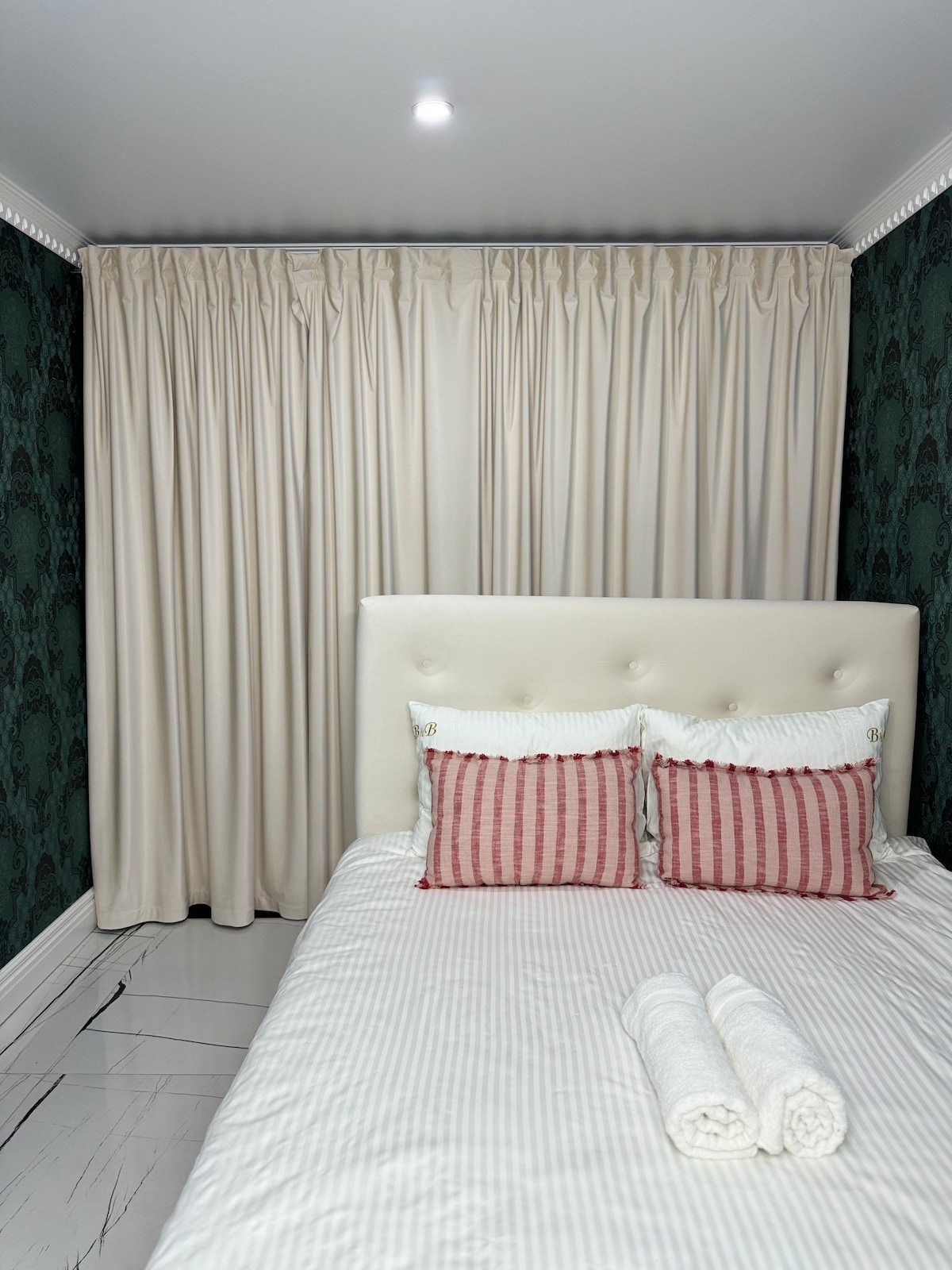 A well-appointed sleeping area features a soft, cream-colored headboard with two decorative pillows in a muted red stripe pattern. Fresh towels are neatly arranged at the foot of the spacious bed, and light-toned curtains frame a window, complementing the dark green patterned wall.
