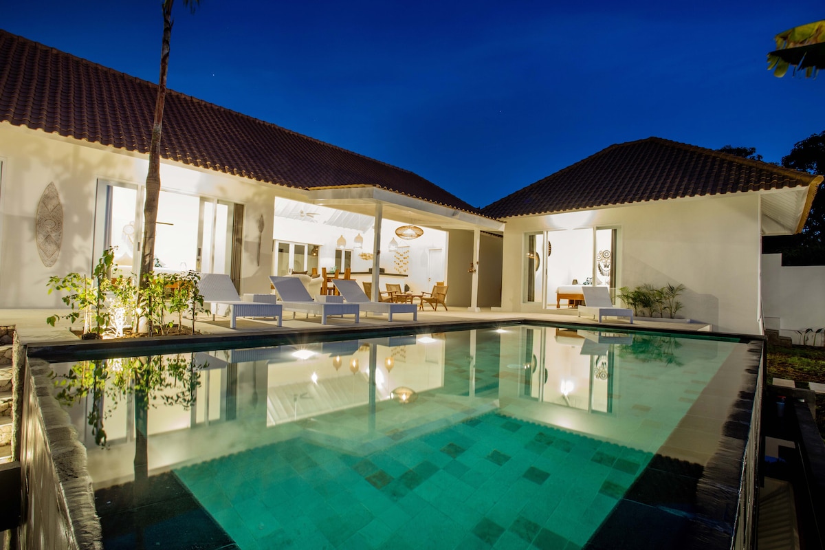 An inviting villa is set against a twilight sky, featuring a modern design and an expansive infinity pool. Lounge chairs are positioned by the water's edge, reflecting soft illumination. Glass doors connect the outdoor space to an elegantly furnished living area, enhancing the seamless indoor-outdoor experience.