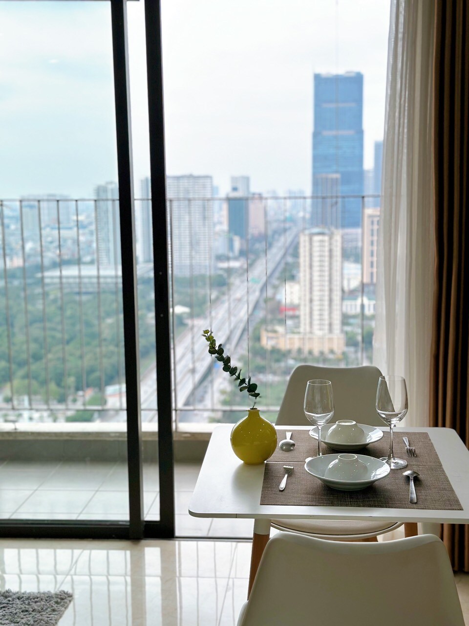A dining table is set for two, featuring a grey placemat, dishware, and glassware. A bright yellow vase holds a single flower. Large windows frame a city view, showcasing towering buildings and green areas, with natural light flooding the room.