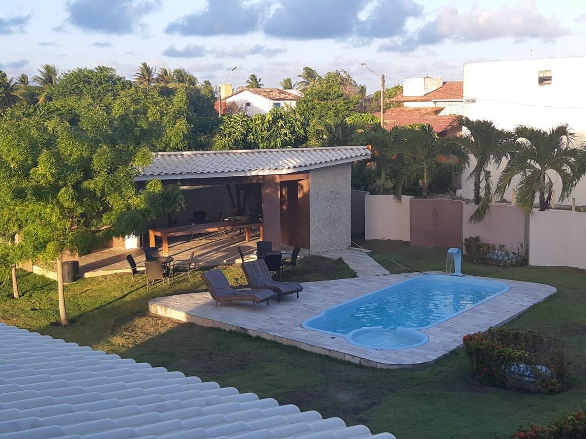 An outdoor area is visible, featuring a rectangular pool surrounded by lounge chairs. A shaded structure with seating is located nearby, complemented by green grass and palm trees. The scene is framed by a low wall, providing privacy and a sense of openness.