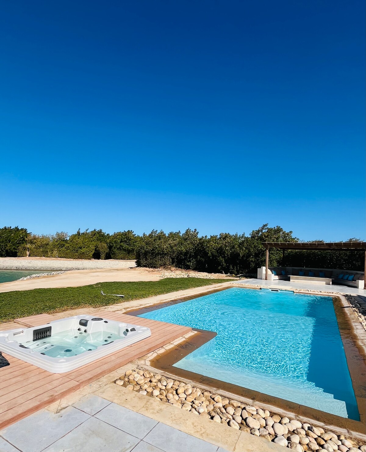 A private pool area is showcased, featuring a sparkling swimming pool with a relaxing hot tub adjacent to it. The surrounding landscaped gardens are visible, set against a clear blue sky, creating a serene outdoor space perfect for leisure and enjoyment.