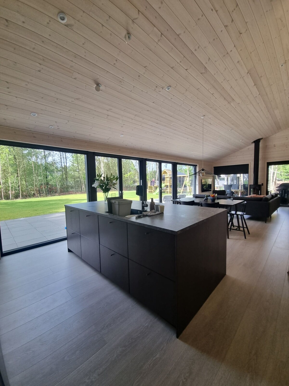 A modern kitchen is featured with a large island, dark cabinetry, and sleek appliances. Expansive glass doors invite natural light and offer views of the outdoor area. A dining table and black chairs complement the open space, enhancing the sense of connectivity between the areas.