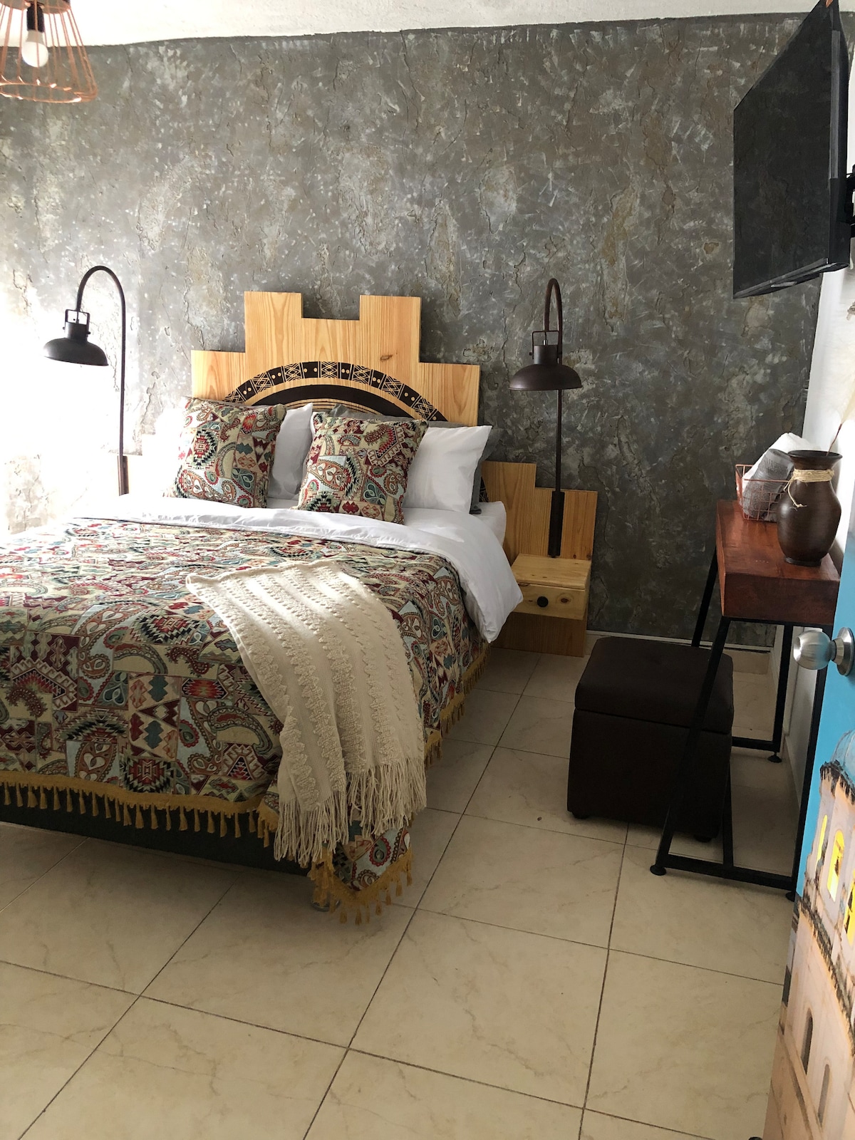 A cozy bedroom features a spacious bed with a colorful patterned blanket and decorative pillows. Unique wooden headboard design is complemented by warm lighting from two wall-mounted lamps. A small bedside table with a drawer and a dark ottoman provide additional comfort, while a television is mounted on the wall.