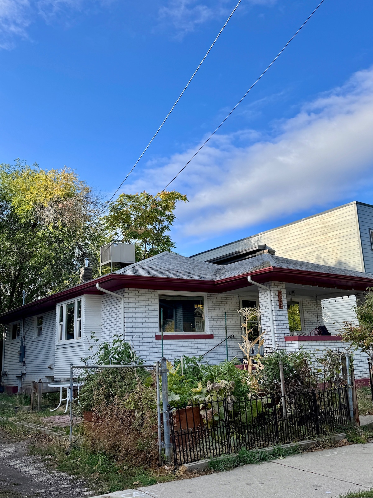 The exterior of a single-story house is shown, featuring a sloped roof and well-maintained landscaping. A front porch with seating is visible, alongside a fenced garden area filled with plants. The surrounding neighborhood is displayed under a partly cloudy blue sky.