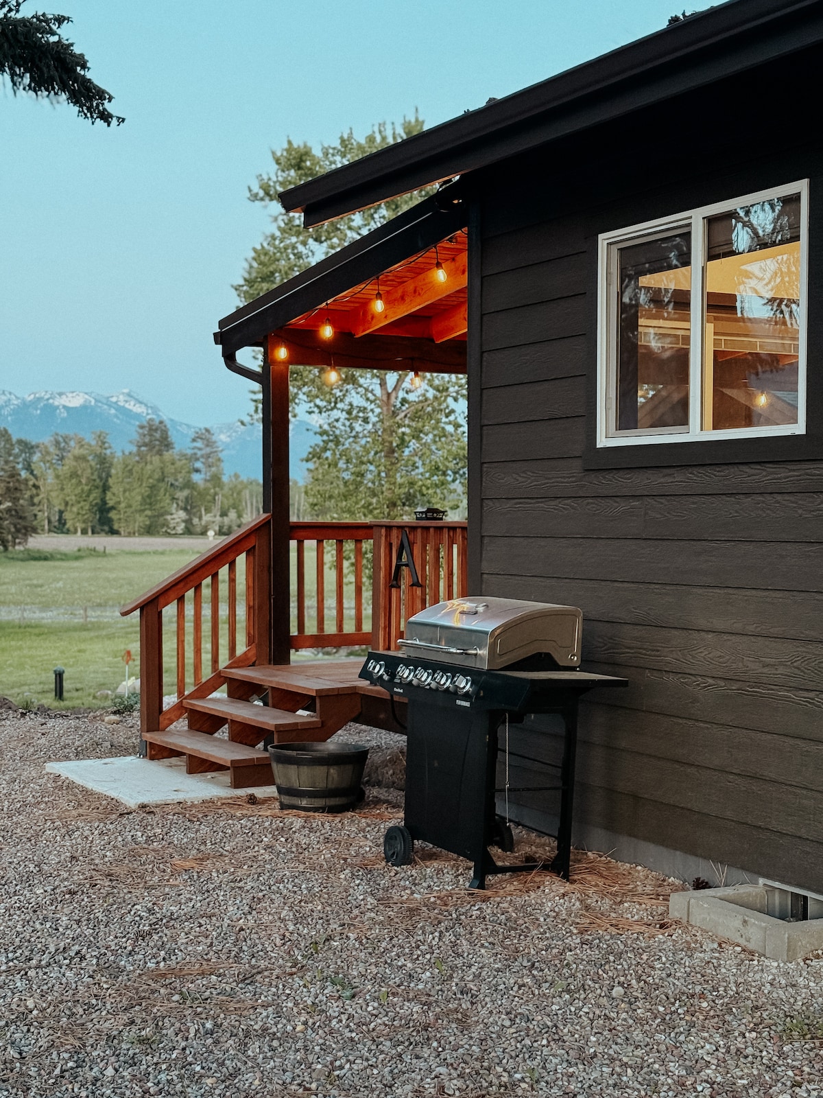 A cabin exterior is illuminated by warm string lights, highlighting the wooden steps and a gas grill placed nearby on a gravel pathway. The surrounding landscape features open fields and distant mountains, creating a serene outdoor setting.