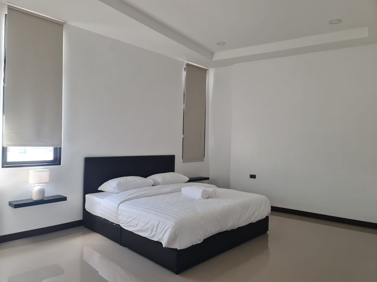 A spacious bedroom is presented with a large bed featuring crisp white linens. Two neutral-toned window coverings allow natural light to enter the room. A small bedside table holds a lamp, and the polished floor adds to the clean aesthetic.