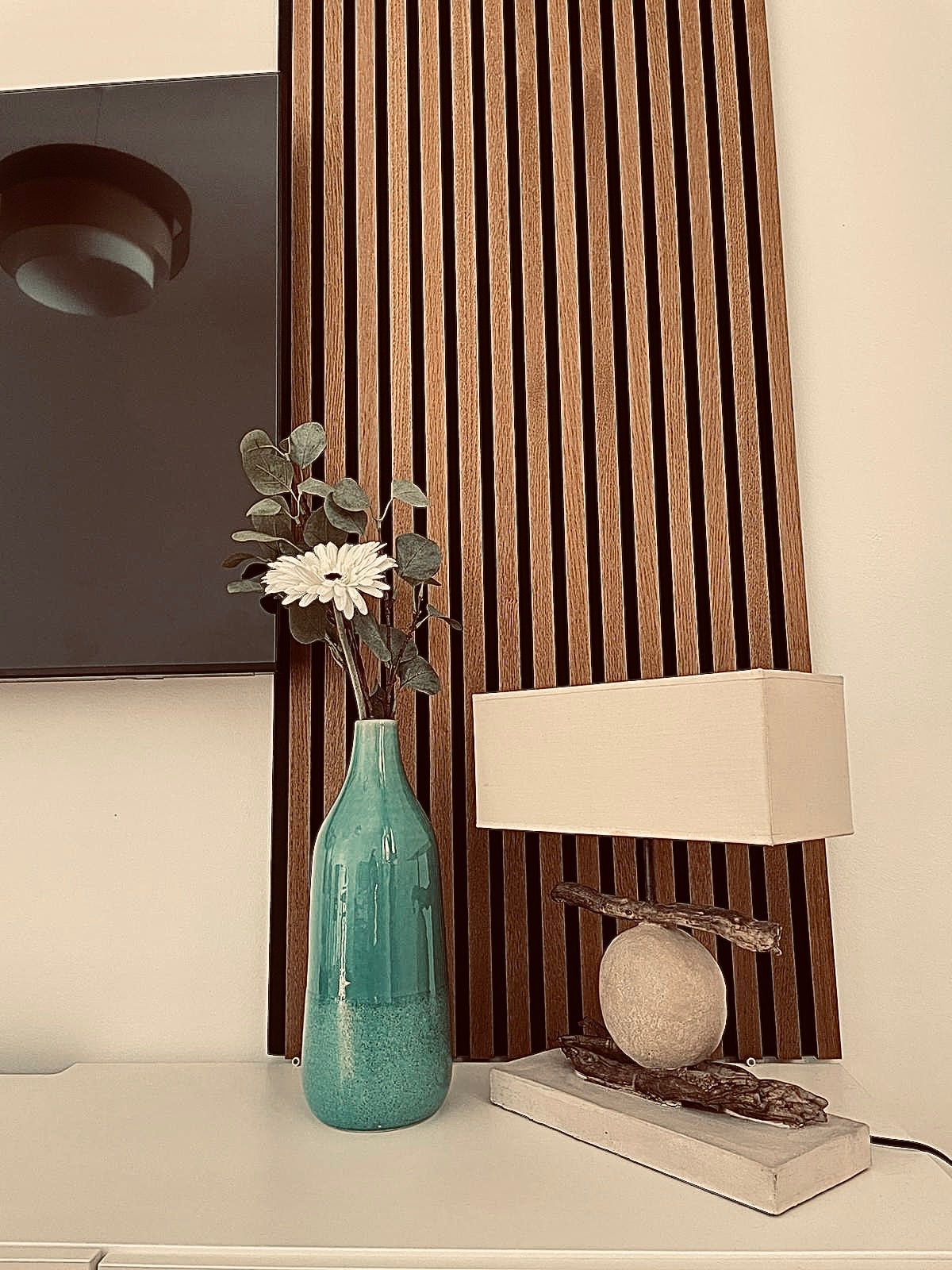 A decorative corner showcases a tall, teal vase with greenery and white flowers. Beside it, a lamp with a simple shade is positioned on a light-colored table, which is complemented by a wood-paneled backdrop adding texture to the space.