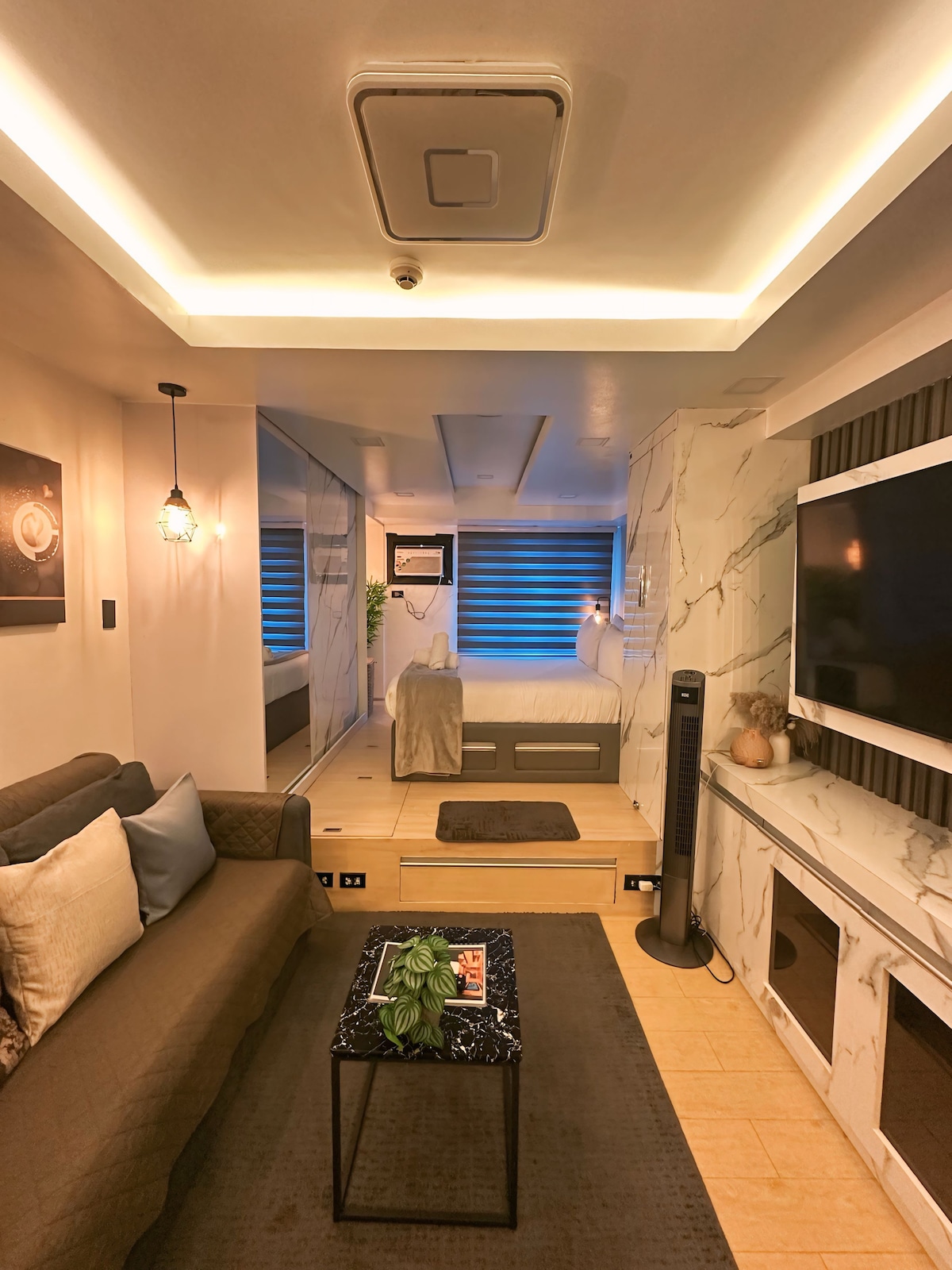 The living area is designed with a sleek appearance, featuring a cozy sofa and a coffee table adorned with greenery. A large TV is mounted on the wall, while a well-defined sleeping area is accessible in the background, illuminated by ambient lighting.