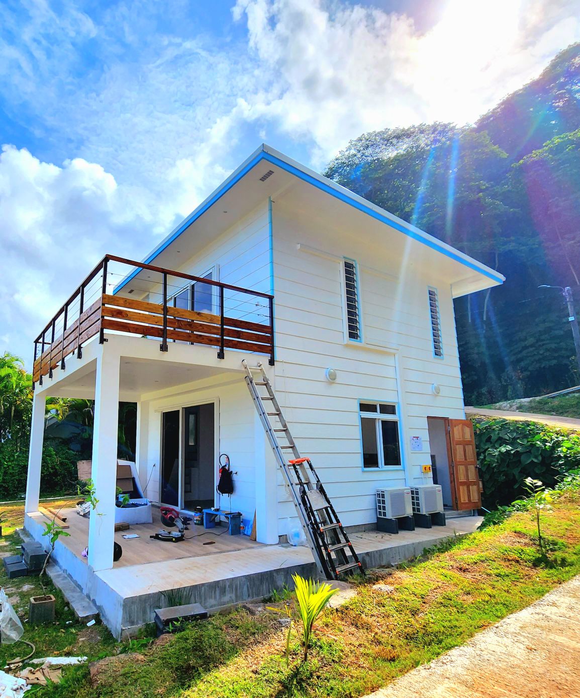 A two-story duplex with a light-colored exterior features a balcony on the upper level. An aluminum ladder is positioned against the building, and air conditioning units are mounted on the side. Surrounding greenery complements the structure, with a pathway leading to the entrance.