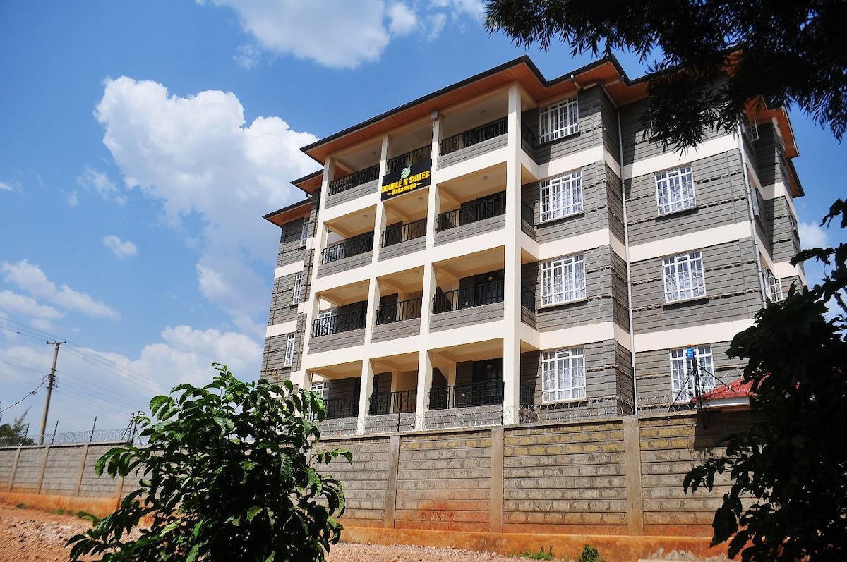 A multi-story building is shown in a sunny setting, featuring a modern architectural design with balconies and large windows. A landscaped area with green foliage is visible in the foreground, highlighting the property's outdoor environment.