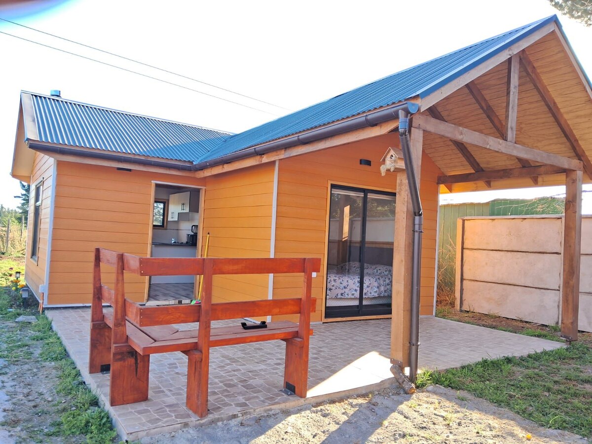 An inviting cabin exterior is shown, featuring wooden elements and a sloped roof. A sturdy bench offers a place to relax. Large glass doors provide a view into the interior, with light filtering through. The surrounding area is open and grassy.