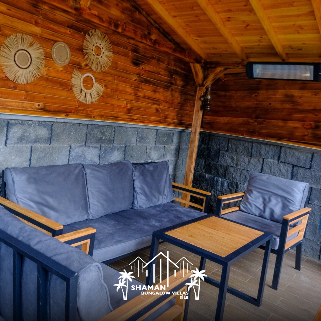 An outdoor seating area features a comfortable gray sofa with wooden armrests, complemented by two matching chairs. A wooden table sits in the center. The surrounding walls are finished in stone, and decorative wall hangings enhance the rustic ambiance.