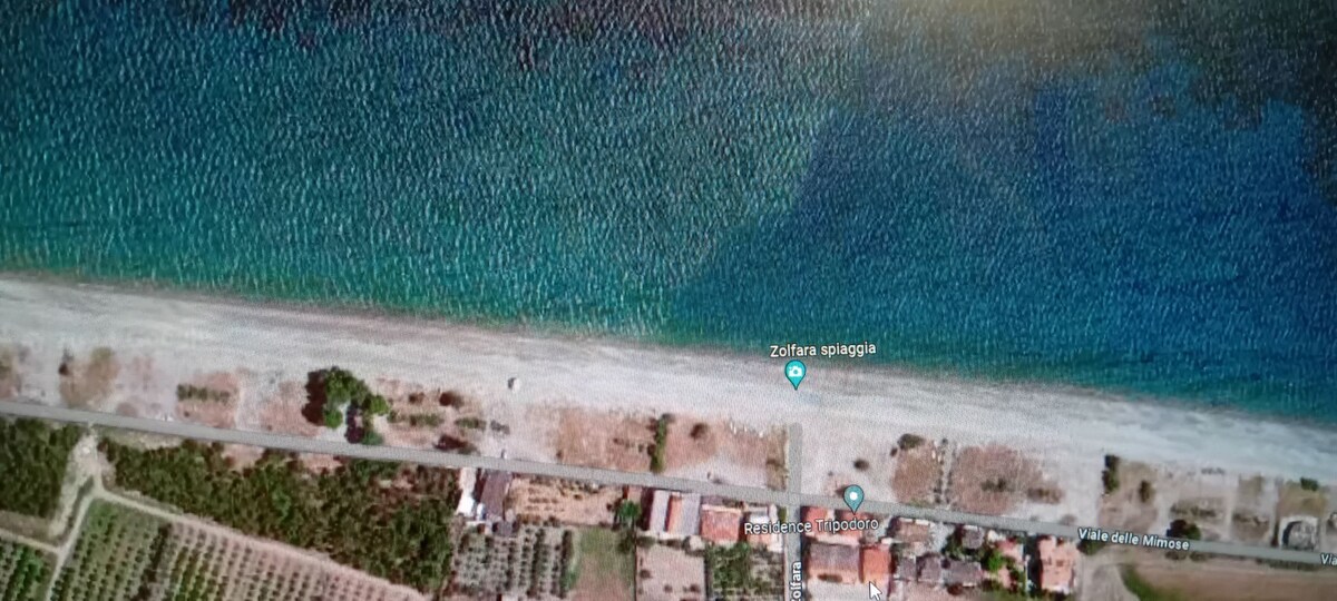 An aerial view captures a peaceful beach area marked by soft sandy shores and clear blue waters. The shoreline is bordered by a road and greenery, while surrounding properties are visible in the landscape, blending naturally into the serene coastal setting.