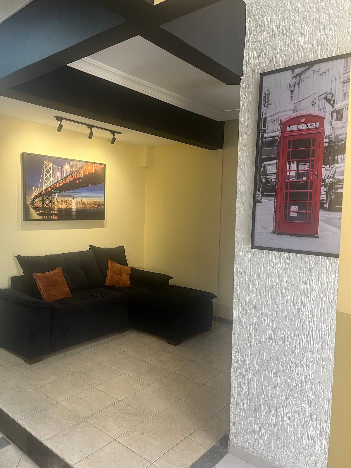 A seating area features a black sectional sofa adorned with contrasting orange cushions. A large framed photograph of a cityscape with a bridge enhances the space, while a framed image of a red telephone booth is positioned on the adjacent wall.