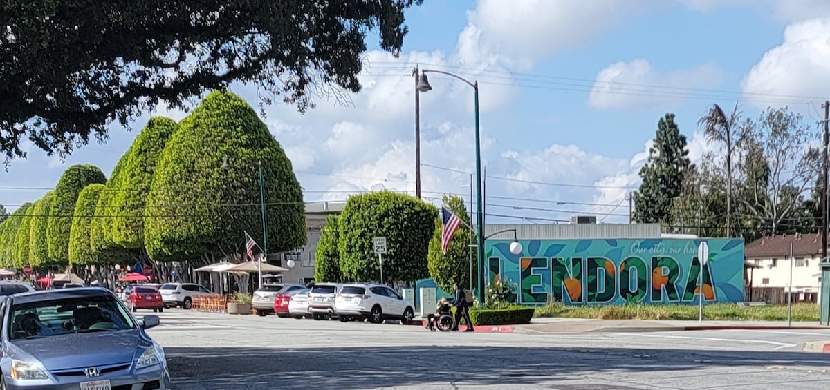 A colorful mural spelling 'Glendora' is displayed on a wall, surrounded by neatly trimmed trees and parked vehicles. The scene is set on a sunny day, with fluffy clouds in the sky, capturing the vibrant atmosphere of the area.