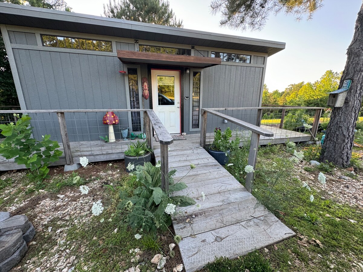 The entrance to a stylish cabin features a wooden ramp leading to a door framed by large windows. A bright red mushroom statue and planter boxes with greenery decorate the front yard, complemented by scattered white flowers and a pathway of rustic planks.