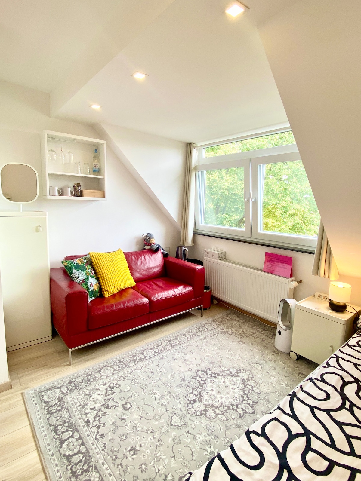 A cozy room features a vibrant red sofa adorned with colorful cushions. Natural light spills through a large window, illuminating the space. A decorative shelf is mounted on the wall, displaying various items. A patterned rug adds texture to the wooden floor.