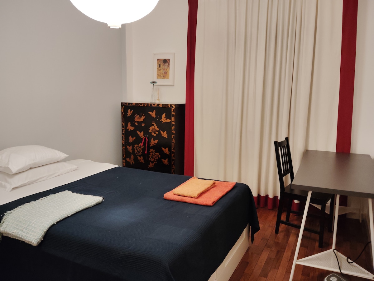 A cozy bedroom features a spacious bed covered with a dark blanket. Two neatly arranged towels are placed at the foot of the bed. A simple desk and chair are positioned against the wall, while a wardrobe with patterned design adds character to the room.