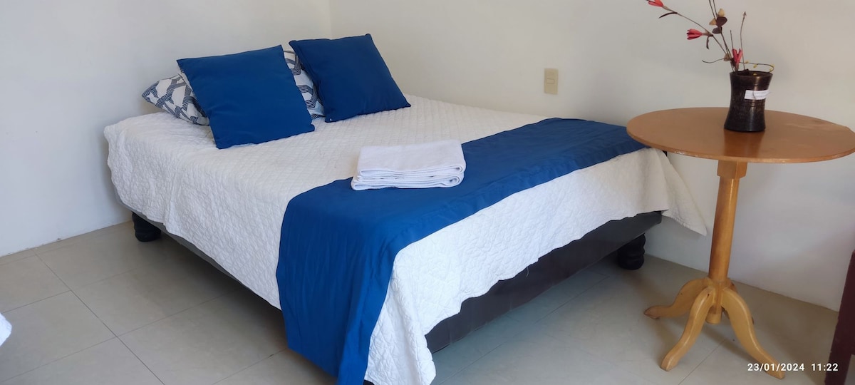 A spacious bed with a white quilted coverlet is flanked by two blue decorative pillows. A small wooden table stands nearby with a dark-colored vase containing artificial flowers. The flooring is light-colored tile, enhancing the room's serene ambiance.