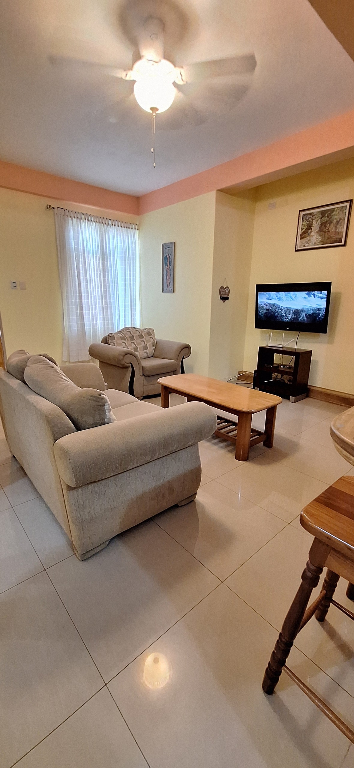 The living area features a light-colored sofa and an armchair, arranged around a wooden coffee table. A television stands on a small stand. Natural light filters through sheer curtains, illuminating the space with a soft glow.