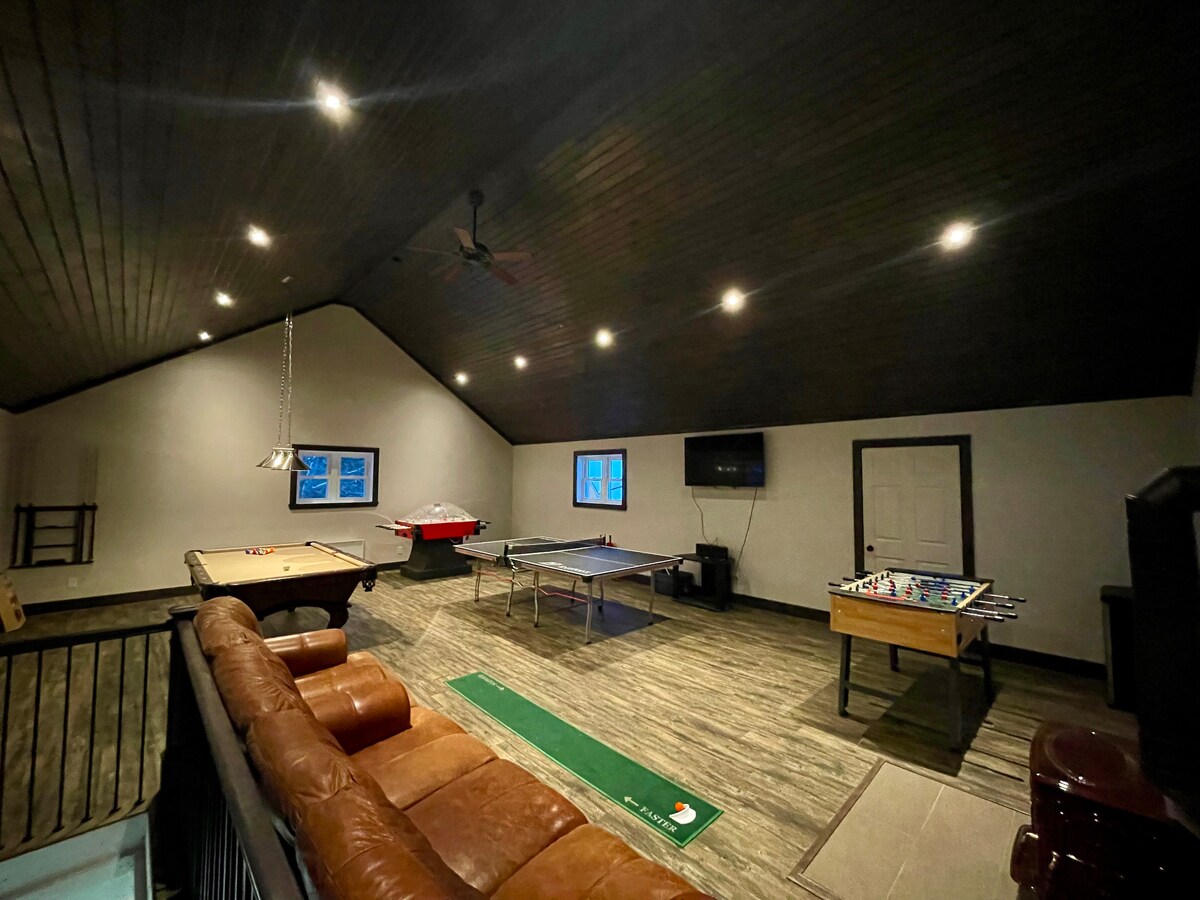 A spacious recreational area is revealed, featuring a dark wood-paneled ceiling and well-lit spaces. Game tables, including pool and foosball, line the room. A comfortable sofa sits in one corner, complemented by a green area rug, while a large flat-screen TV is mounted on the wall.