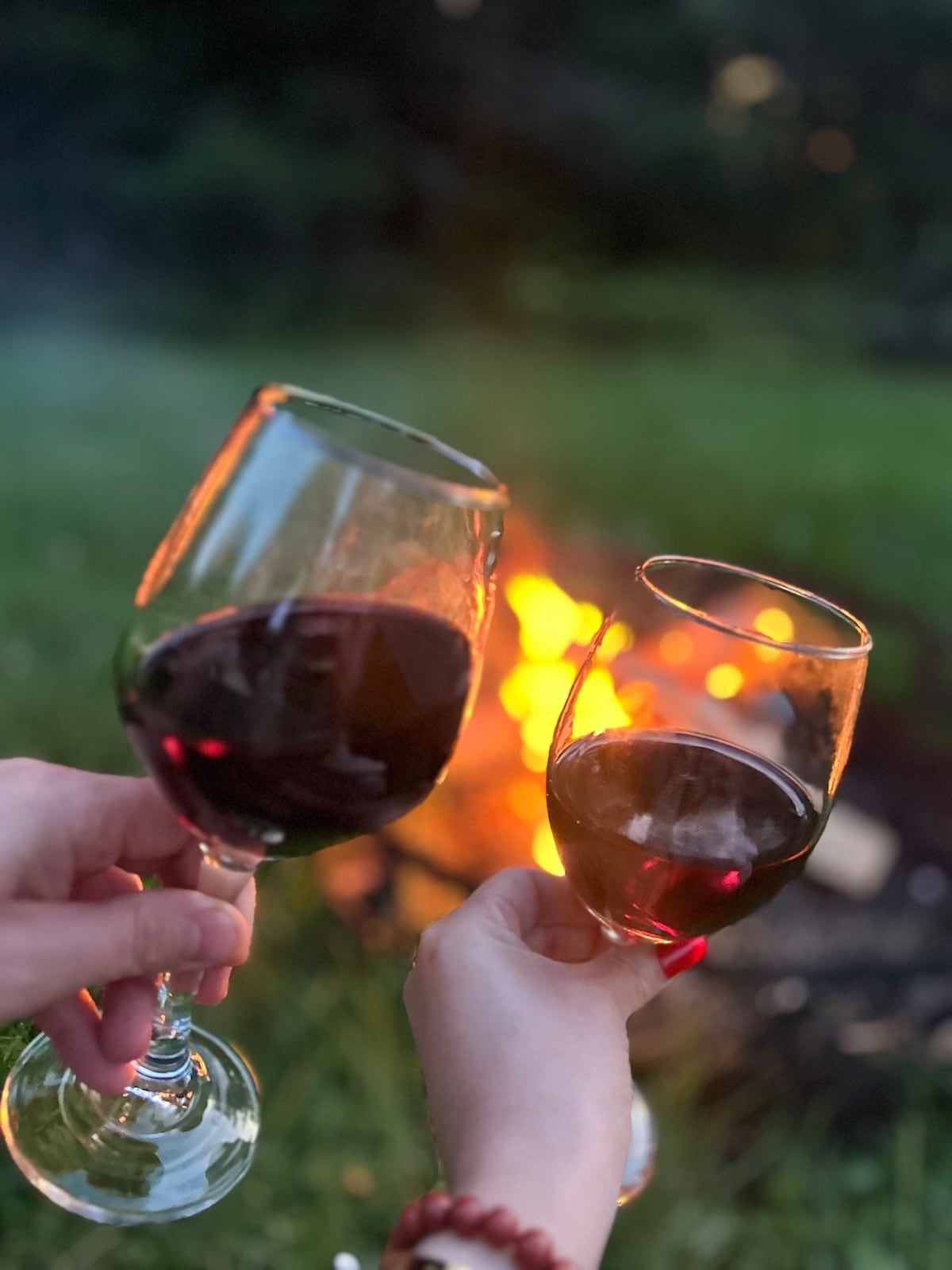 Two glasses of red wine are raised in a toast against the backdrop of a glowing fire pit. Soft flames emit a warm light, illuminating the grassy area surrounding the fire. The image captures a cozy outdoor setting ideal for relaxation.
