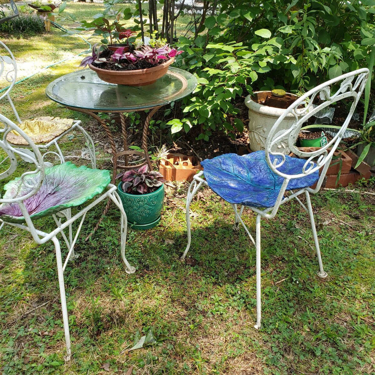 A small outdoor seating area is surrounded by lush greenery. Two wrought-iron chairs with colorful leaf-patterned cushions flank a circular glass table. A potted plant sits on the table, adding a touch of nature to the peaceful setting.