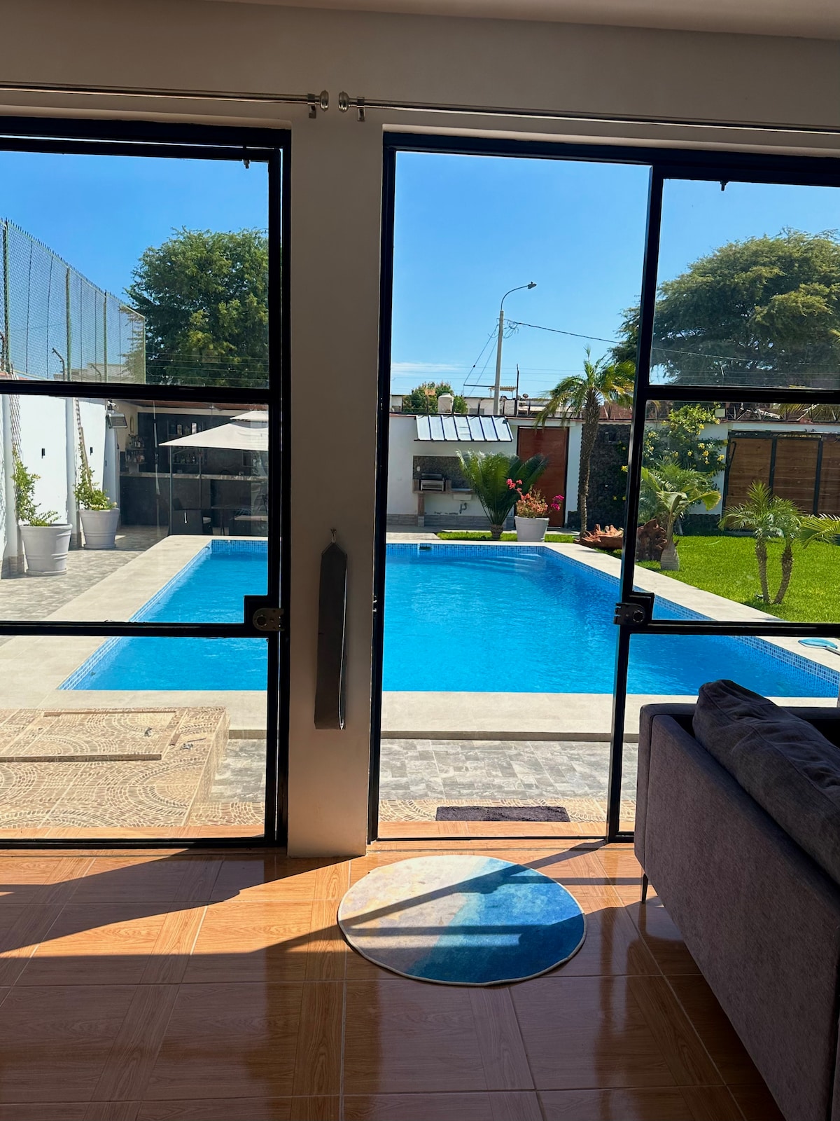 Natural light floods the interior space through expansive glass doors that open to a private pool area. The view includes a well-maintained swimming pool bordered by a green space, as well as a covered outdoor seating area. The ambiance reflects a seamless connection between indoors and outdoors.