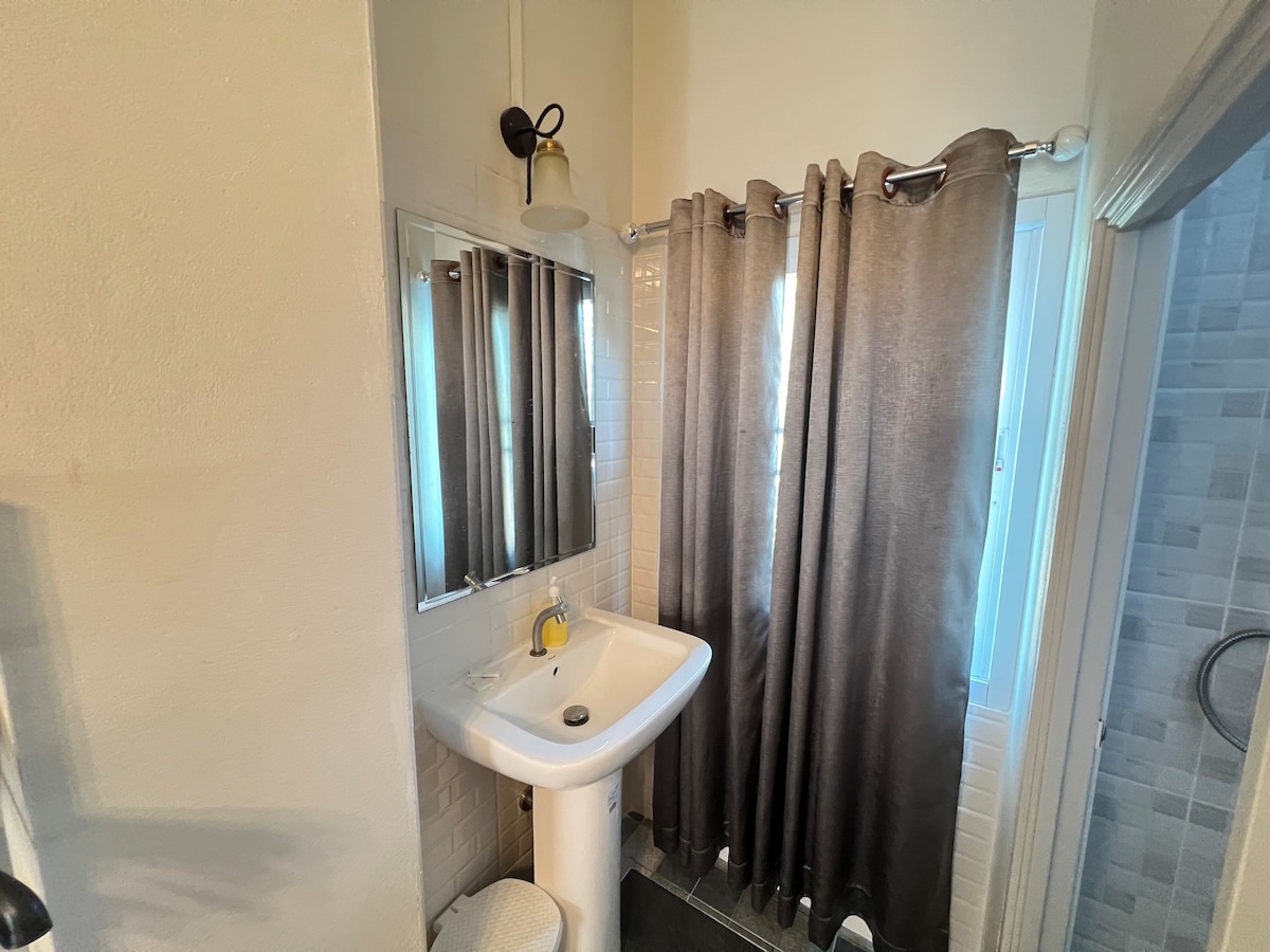 A simple bathroom is captured, featuring a white sink mounted with a gold faucet. A large mirror reflects the space, complemented by gray curtains that frame a window. Light enters the room, enhancing the bright, clean feel of the area.