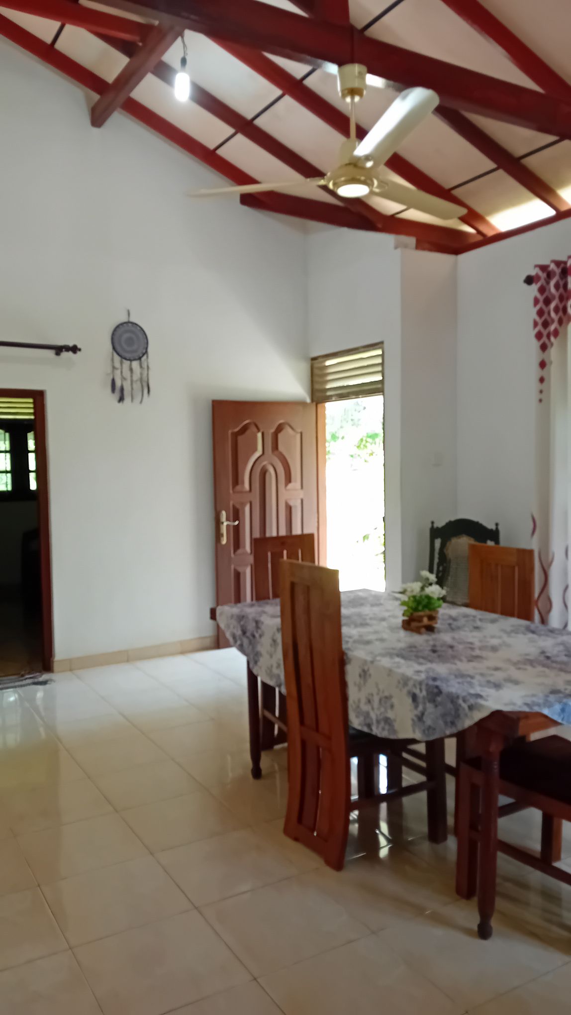 A dining area is presented with a large table surrounded by wooden chairs. Natural light enters through the adjacent window, highlighting the smooth tiled floor. A dreamcatcher hangs near the ceiling, complementing the open and airy feel of the space.