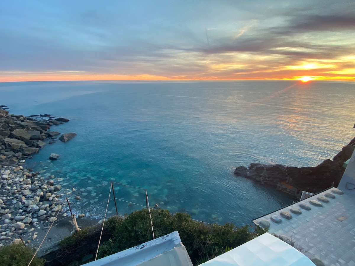 A serene coastal view captures the tranquil sea at sunset, with soft reflections of orange and pink hues on the water. Rocky formations line the shoreline, while clear waters reveal a smooth sandy bottom, creating a peaceful atmosphere.