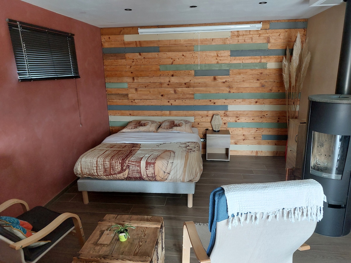 The spacious main area includes a comfortable bed with decorative bedding, flanked by two nightstands. A seating area features a chair and an accent chair, while wooden accents and a charming wood stove enhance the ambiance. Light enters through a window with dark blinds.
