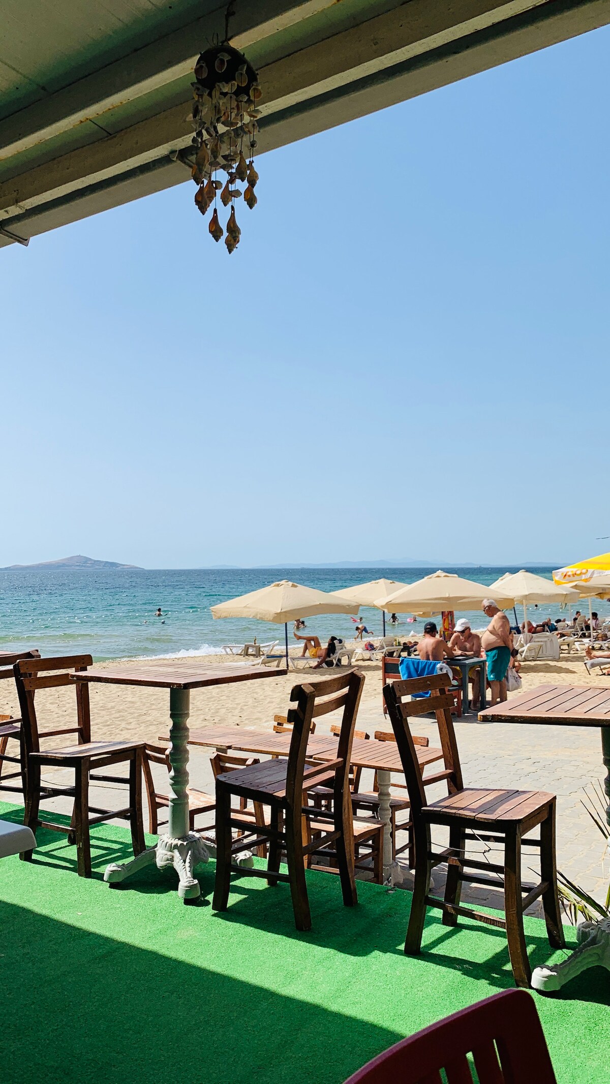 A beachfront scene shows a sandy shore with several umbrellas providing shade to beachgoers. Wooden chairs and tables are positioned on a green mat, offering a view of the ocean and distant mountains under a clear blue sky.