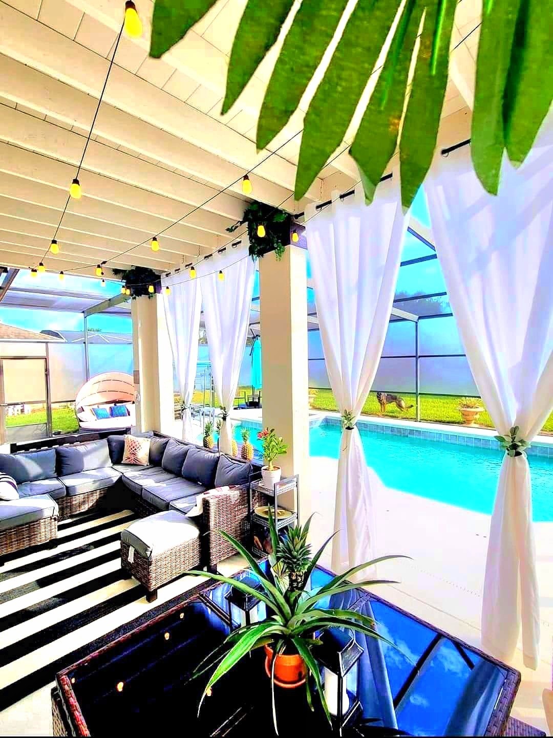 A covered outdoor seating area features a dark wicker sofa arranged around a glass-top table. Sheer white curtains hang beside the pool, which is visible through the screened enclosure. Soft lighting is provided by string lights above, and a lush green plant adds a natural touch.