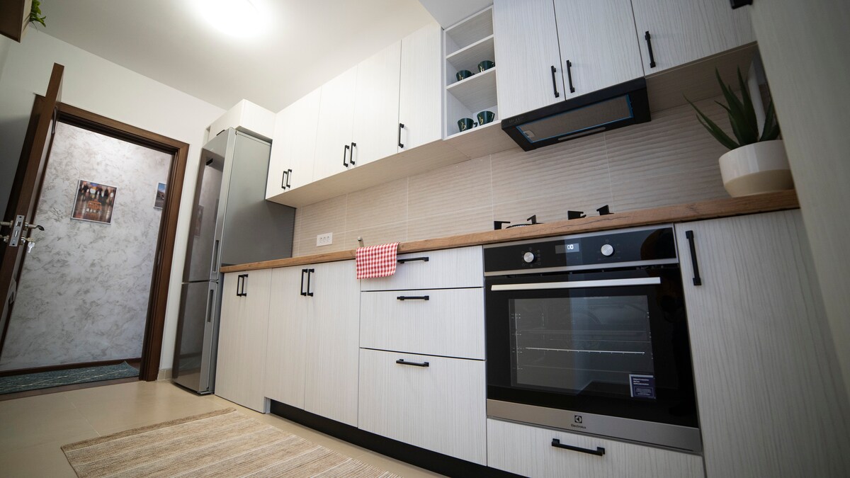 A modern kitchen is featured with sleek cabinetry and a wooden countertop. Stainless steel appliances include an oven and refrigerator. Open shelving displays green mugs, while a potted plant adds a touch of nature. A striped towel is placed on the countertop, enhancing functionality.