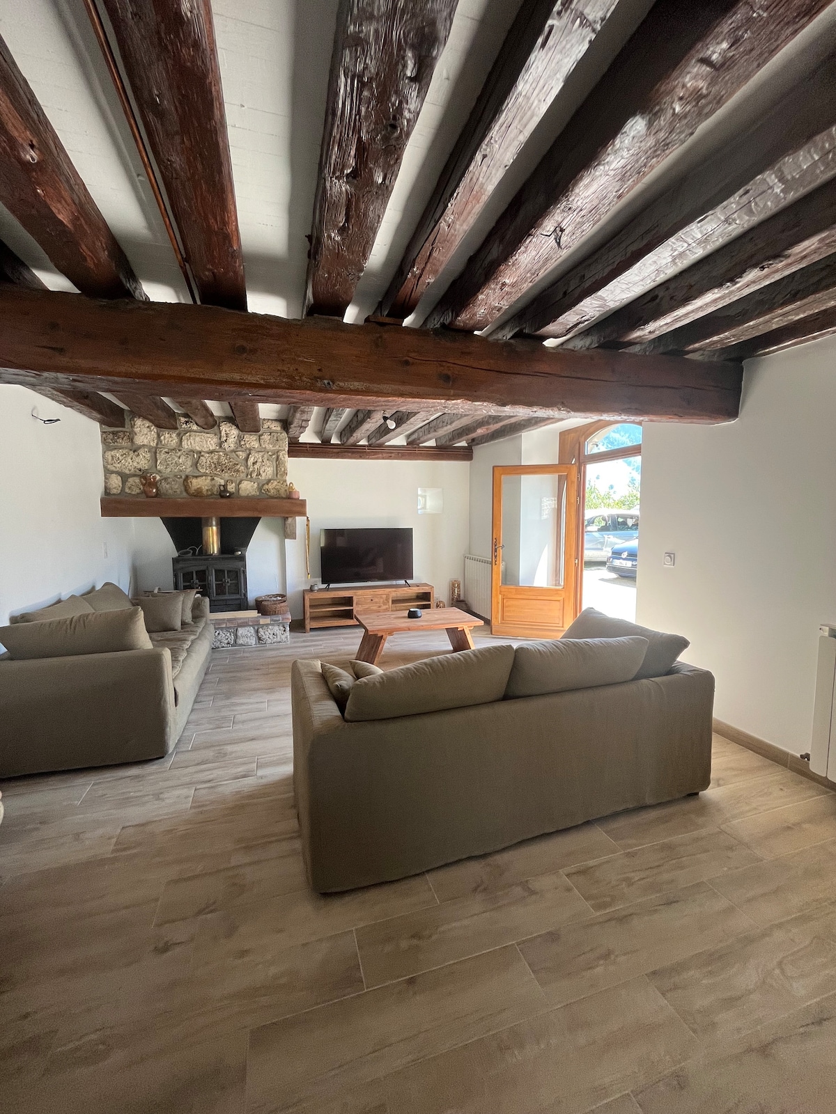 The living area features exposed wooden beams and a stylish decorative fireplace. Two comfortable sofas are positioned for relaxation, complemented by a wooden coffee table and a television. Large doors provide natural light and a view of the exterior.