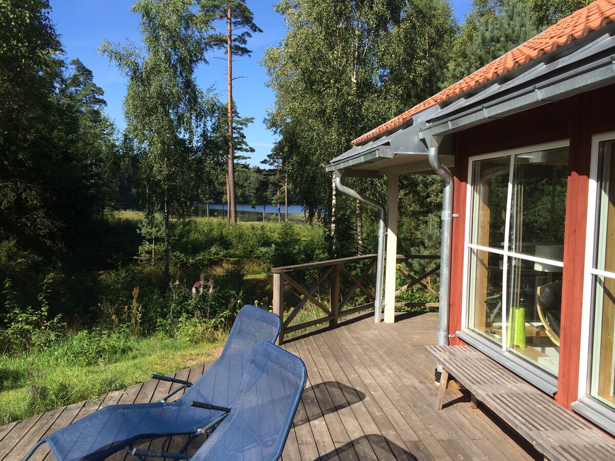 A wooden deck extends from the cabin, featuring two lounge chairs positioned for relaxation. The view beyond includes a serene lake, framed by lush green trees and clear blue skies, creating a peaceful natural setting.