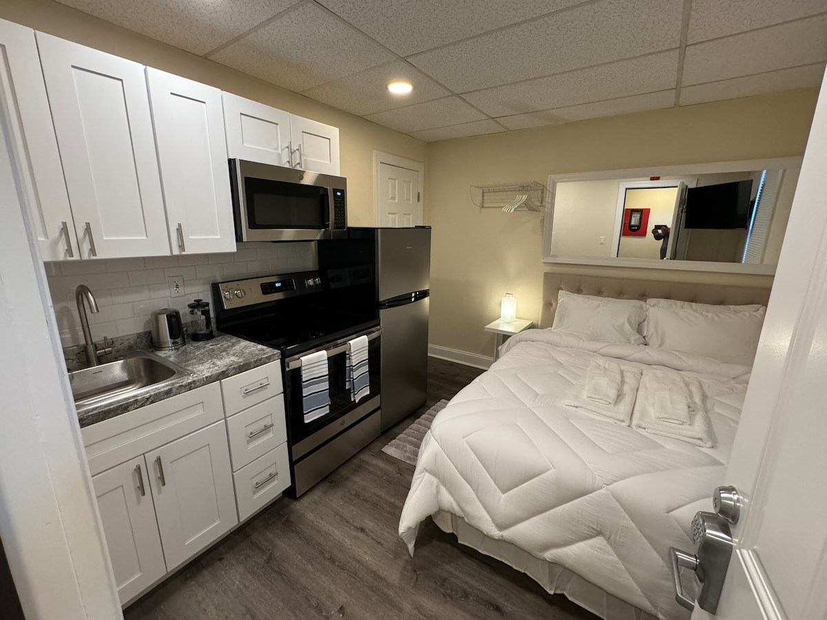 A compact studio apartment features a cozy sleeping area with a spacious bed covered in white bedding. A kitchenette with modern appliances, including a microwave and a refrigerator, is visible. Light from a lamp creates a soft glow near the bed.