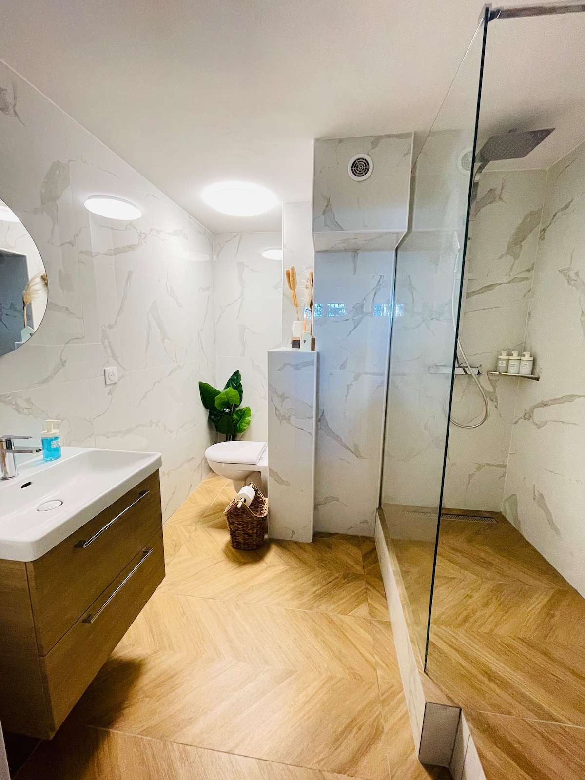 A modern bathroom is presented with marble-patterned walls and warm wooden flooring. A sleek, white vanity sits beneath a round mirror, alongside a glass-enclosed shower featuring a rain showerhead. Fresh towels are neatly placed on the floor, complemented by decorative greenery.