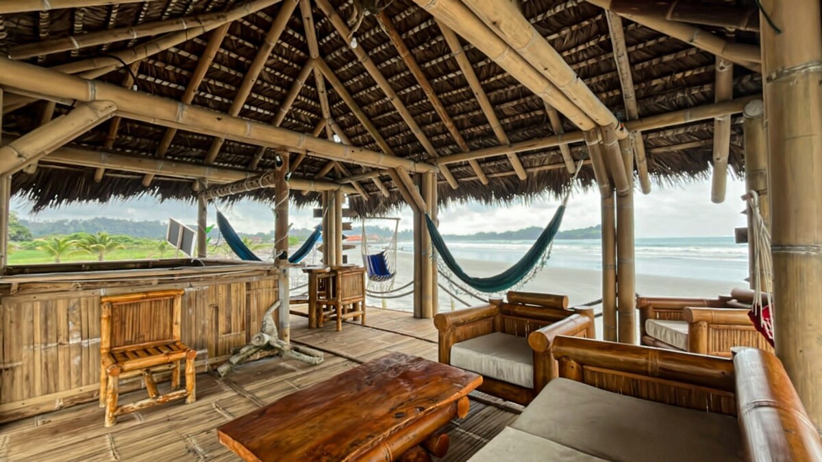 A covered outdoor lounge area is constructed with bamboo and thatch, featuring comfortable seating arrangements and a wooden coffee table. Hammocks are hung nearby, offering a relaxed spot. The backdrop showcases the beach and ocean, with soft natural lighting filtering through.