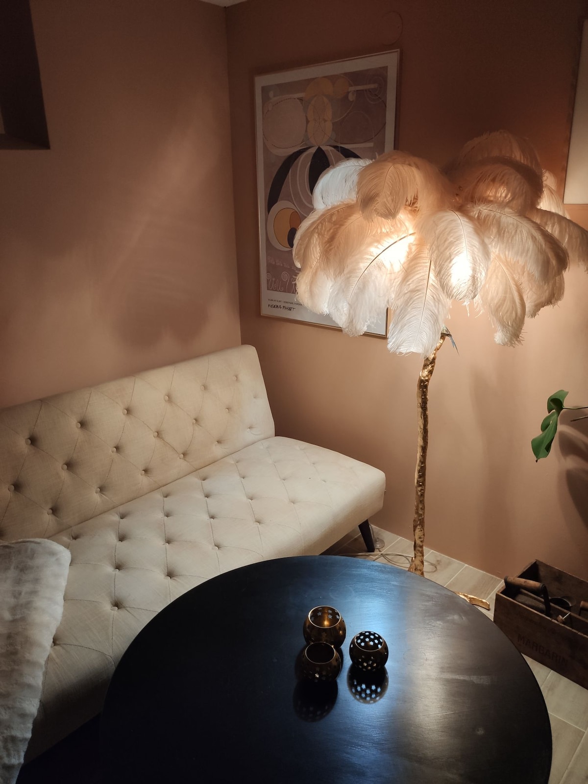 A charming seating area is displayed, featuring a light-colored tufted futon adjacent to a tall, decorative floor lamp with feather-like elements. A round black table is positioned in front, adorned with three decorative candle holders. Soft lighting enhances the warm tones of the walls.