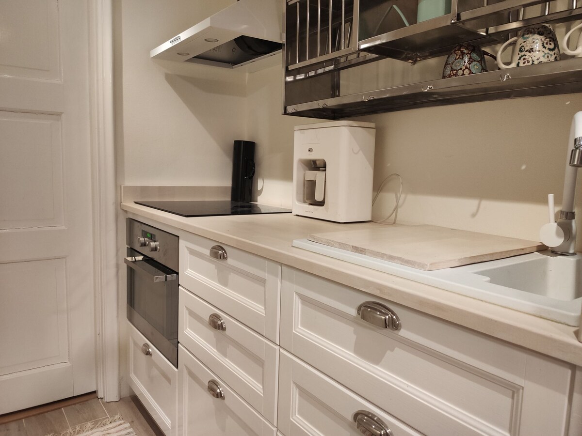 A modern kitchen features white cabinetry with sleek silver hardware. An induction cooktop and an oven are integrated into the counter space. Above, a shelf displays decorative mugs. A coffee maker is positioned on the counter, complementing the contemporary design.