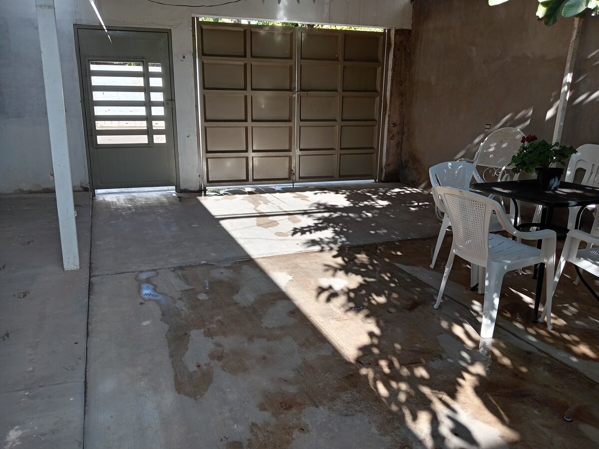A shaded outdoor area features a concrete floor with light and shadow patterns created by overhead foliage. A patio table is accompanied by white chairs, while a garage door is visible at the back, contributing to a functional and open space.